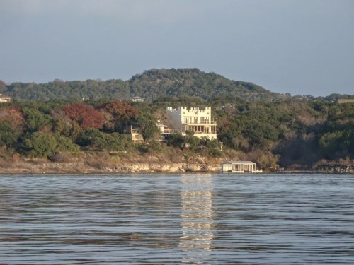 A large house on a hillside overlooking a calm lake, surrounded by trees and distant hills under a clear sky.