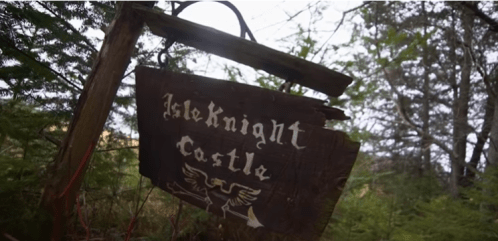 Weathered wooden sign for "Isleknight Castle" hanging in a forested area, surrounded by trees.