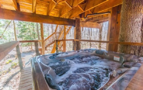 A wooden hot tub under a rustic cabin, surrounded by trees and a staircase leading to the upper level.