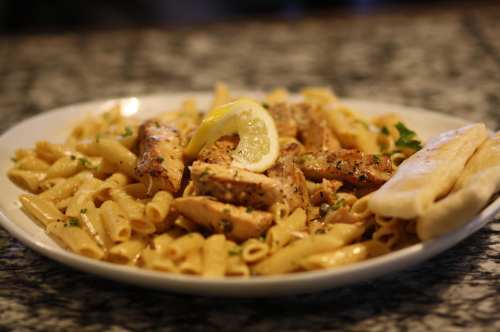 A plate of pasta with grilled chicken, lemon slices, and a side of bread, garnished with herbs on a granite surface.