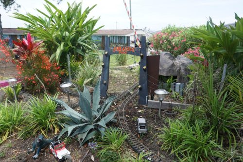 A miniature Jurassic Park entrance with toy cars, surrounded by vibrant plants and a rocky feature.