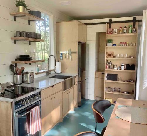 Modern kitchen with wooden cabinets, stainless steel appliances, open shelves, and a dining table. Bright and airy atmosphere.