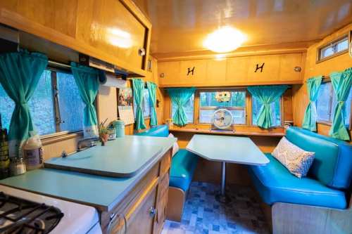 Cozy vintage camper interior with teal curtains, a dining table, and a kitchenette featuring a stove and sink.