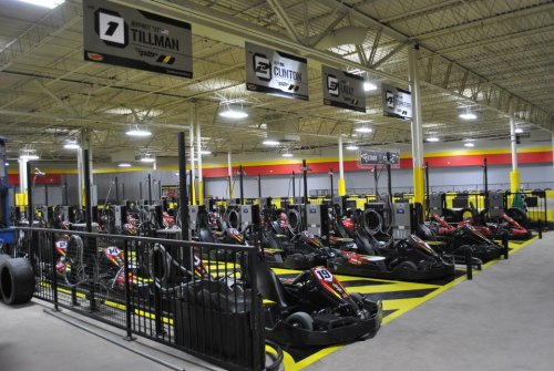 Indoor go-kart racing track with multiple karts lined up, surrounded by safety barriers and overhead signage.