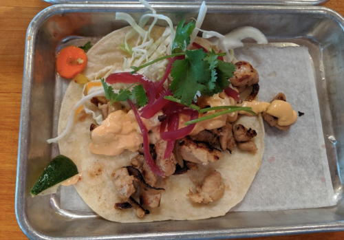 A taco on a metal tray, filled with grilled chicken, cabbage, pickled onions, cilantro, and a creamy sauce.