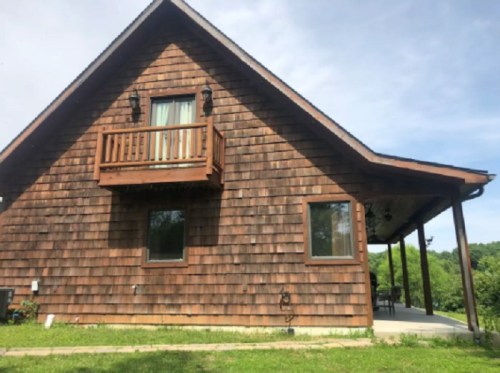 A wooden cabin with a sloped roof, balcony, and large windows, surrounded by green grass and trees.