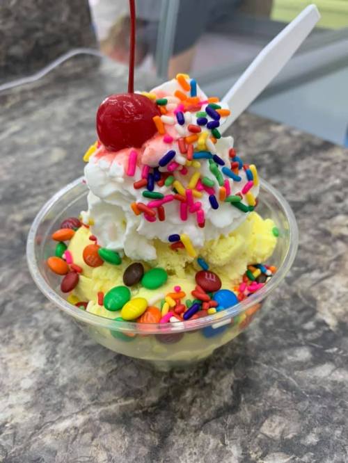 A colorful ice cream sundae topped with whipped cream, sprinkles, a cherry, and chocolate candies in a clear cup.