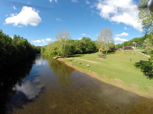 A serene river scene with green trees, a grassy bank, and a clear blue sky dotted with clouds.