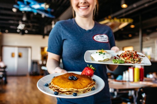 A smiling waitress holds two plates of food: one with pancakes topped with berries and another with a savory dish.