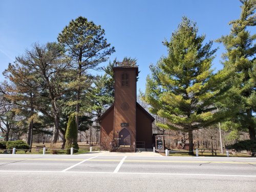 A small brown church surrounded by tall trees on a clear blue sky day, viewed from across the road.