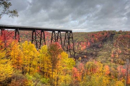 A tall bridge spans a colorful autumn landscape with vibrant trees under a cloudy sky.