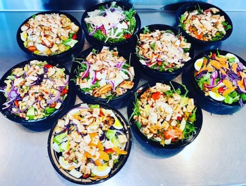 A variety of colorful salads in black bowls, featuring greens, vegetables, grilled chicken, and toppings.