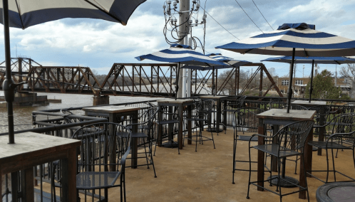 Outdoor seating area with striped umbrellas overlooking a river and a bridge in the background.