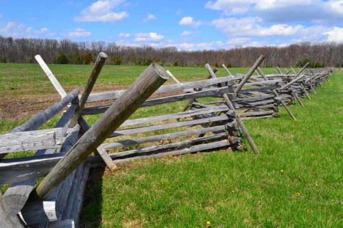 A weathered wooden fence stretches across a green field under a blue sky with fluffy clouds.