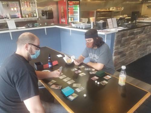 Two people wearing masks play a card game at a restaurant table, with drinks and snacks nearby.