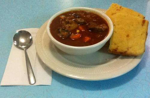 A bowl of hearty beef stew with vegetables, served with a slice of cornbread and a spoon on a blue table.