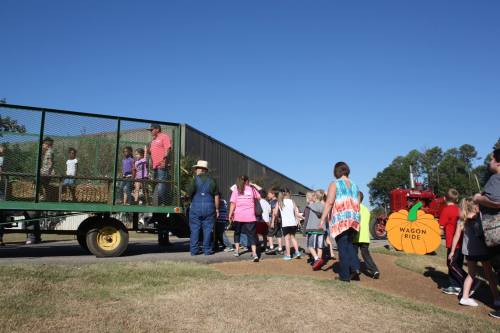 Children and adults line up for a wagon ride, with a tractor and a sign in a sunny outdoor setting.