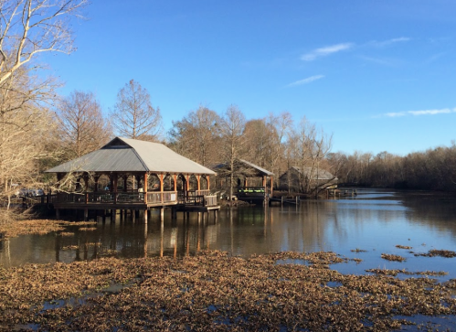 A serene view of wooden pavilions by a calm lake, surrounded by trees and clear blue skies.