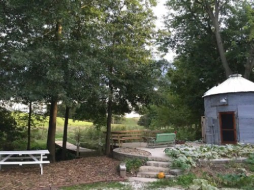 A rustic outdoor space with a round metal structure, picnic table, and lush greenery surrounding a serene landscape.