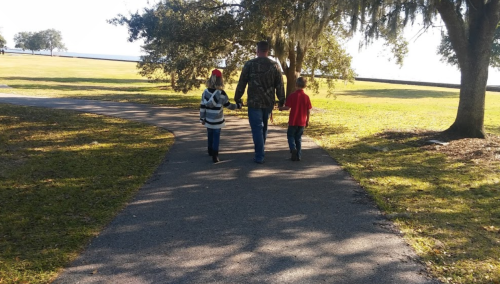 A man walks hand-in-hand with two children along a path in a park, surrounded by trees and open grass.