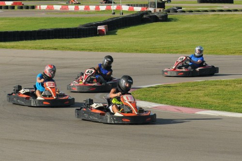 Five go-karts race around a track, with drivers wearing helmets and colorful racing suits, on a sunny day.