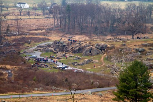 A scenic view of rocky terrain with people gathered, surrounded by trees and fields, under a cloudy sky.