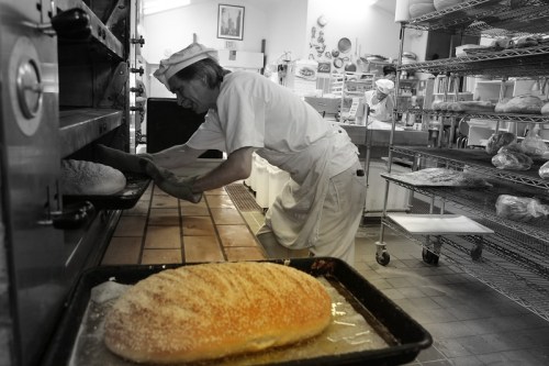 A baker in a white uniform removes freshly baked bread from an oven in a cozy bakery.