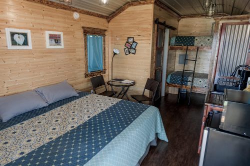 Cozy interior of a wooden cabin featuring a double bed, bunk beds, a small table, and kitchenette.