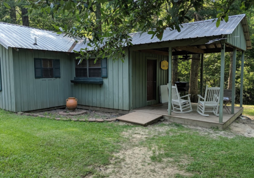 A small green cabin with a porch, rocking chairs, and a clay pot, surrounded by grass and trees.