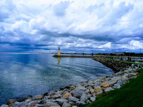 A lighthouse stands on a rocky shore under a cloudy sky, reflecting in calm waters. Green grass lines the pathway.