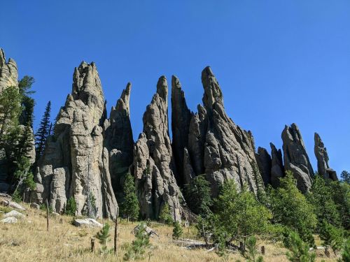 A rugged formation of tall, jagged rock spires rises against a clear blue sky, surrounded by sparse greenery.