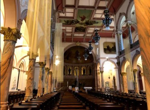 Interior of a church featuring ornate columns, a painted ceiling, and rows of wooden pews. Soft drapes hang from above.