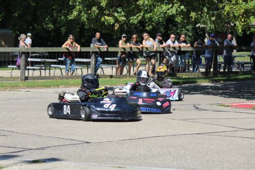 Two go-karts racing on a track, with spectators watching from the sidelines under a sunny sky.