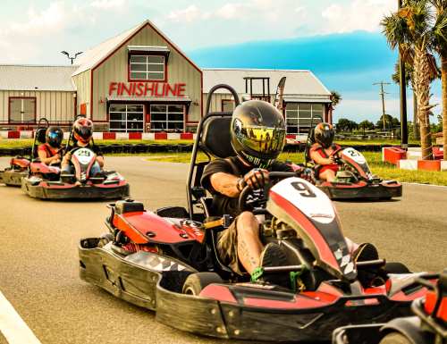 Go-kart racers compete on a track, with a finish line building in the background and palm trees lining the road.