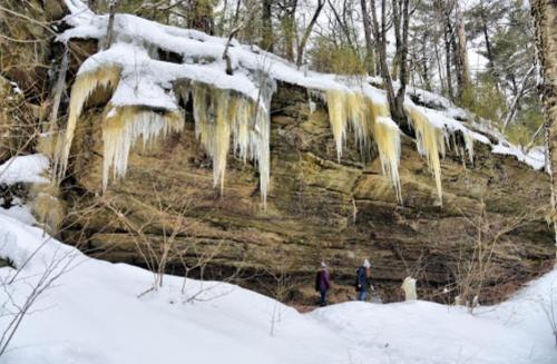 Two hikers walk near a snow-covered cliff with icicles hanging from the rock face in a winter forest.
