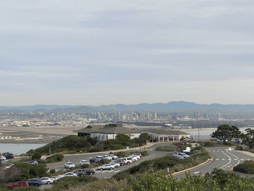 A panoramic view of a city skyline with mountains in the background, featuring a winding road and parked cars in the foreground.
