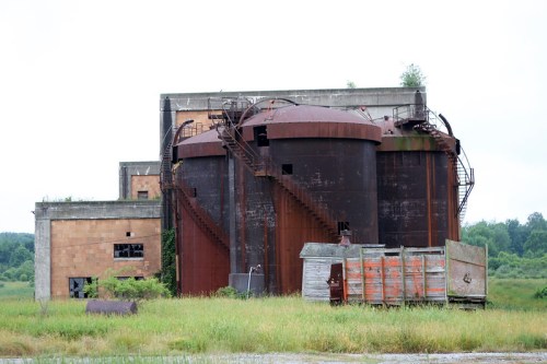 Abandoned industrial site featuring large rusted tanks and a weathered building, surrounded by overgrown grass.