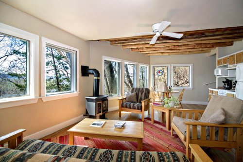 Cozy living area with large windows, wooden beams, a stove, and a dining table, surrounded by nature.
