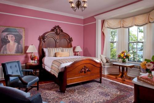 Cozy bedroom with a pink wall, wooden bed, floral decor, and a large window overlooking a scenic view.
