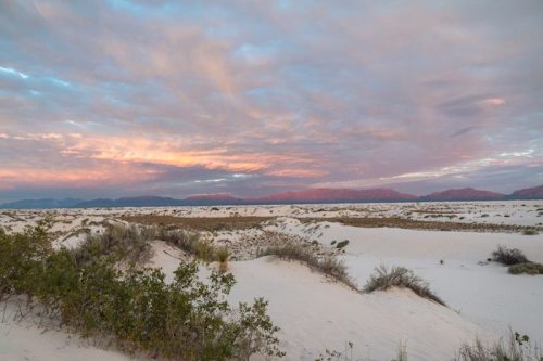 A serene landscape of white sand dunes under a colorful sky at sunset, with sparse vegetation in the foreground.