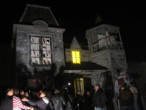 A spooky, old house at night with a glowing yellow window and a crowd of people in front. Shadows create an eerie atmosphere.