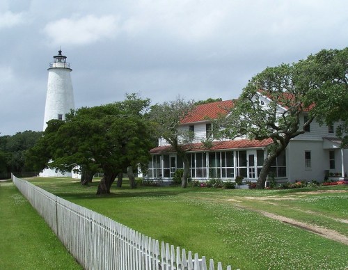 A white lighthouse beside a historic house, surrounded by green grass and trees under a cloudy sky.