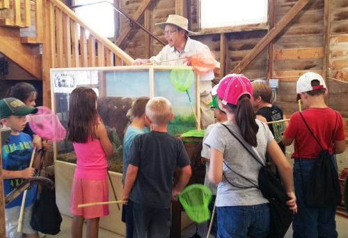 A group of children with nets listens to an instructor by a display tank in a rustic educational setting.