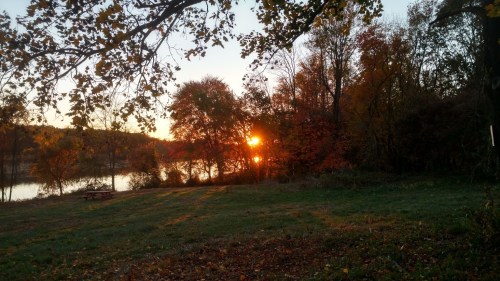 Sunset over a tranquil lake, framed by colorful autumn trees and a grassy area.