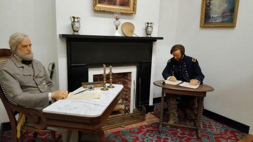 Two life-like figures of historical figures seated at desks, writing, in a room with a fireplace and decorative artwork.