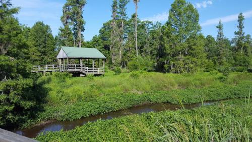A wooden gazebo on stilts overlooks a lush green landscape and a winding stream under a clear blue sky.