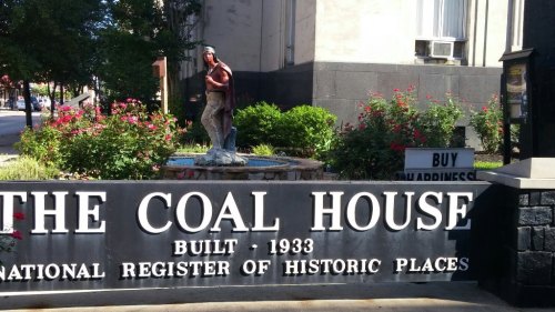 Sign for The Coal House, built in 1933, featuring a statue and surrounded by flowers. Historic site marker included.