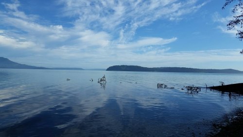 A serene lake scene with calm waters, distant hills, and a blue sky filled with wispy clouds.