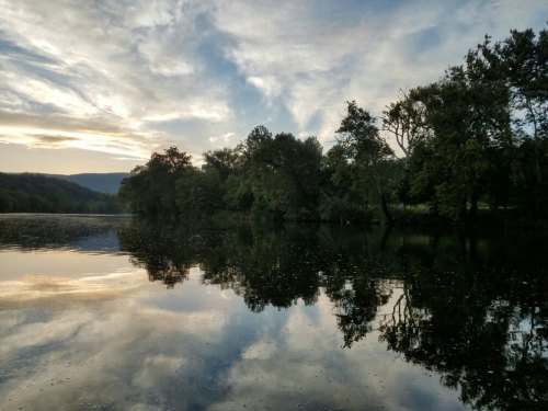 A serene lake at sunset, reflecting trees and clouds in calm waters under a colorful sky.
