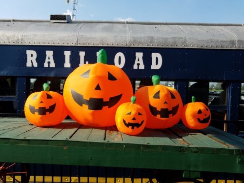 Five inflatable orange pumpkins with black faces displayed on a green platform next to a blue train car.